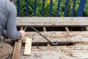 Man working on repairing rotten wood deck boards; male holds a hammer and nearby is a crowbar